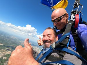 Un homme à la barbe grise sourit à la caméra en faisant des thumbs up pendant que son instructeur pilote leur parachute jaune et bleu.
