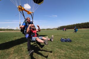 Un client tandem surexcité et souriant lève haut les jambes en prévision de l'atterrissage sur les fesses de son saut en parachute.