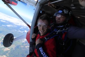 Un jeune homme souriant est à genoux dans la porte de l'avion et s'apprête à vivre un saut en parachute attaché à son instructeur. 