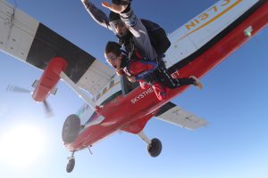 Un invité tandem attaché à sont instructeur de parachute s'élance en chute libre par la porte de côté d'un avion blanc et rouge. 