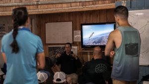 Ryan Blunk, un entraîneur de parachute explique un mouvement de wingsuit à un groupe de parachutiste, aidé d’un vidéo qui défile sur une télévision.