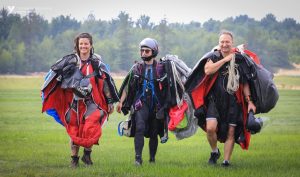 Trois parachutistes vêtus de combinaison ailée, appelée wingsuit marchent dans le champ d’atterrissage avec leur parachute dans les mains. 