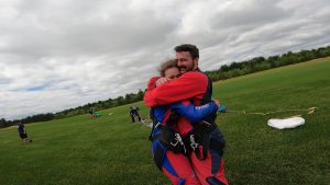 Un homme et une femme habillés de harnais de parachute tandem et de combinaisons de saut rouge s'enlacent après une demande en mariage au moment de l'atterrissage. 