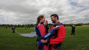 Une passagère de saut en parachute tient un boitier de bague de fiançailles dans sa main, alors que son conjoint enlace sa taille après l'atterrissage de leur sauts en parachute.