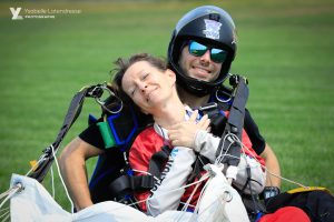 Une maman souriante presse les mains sur son coeur et savoure l'atterrissage de son 1er saut en parachute assise dans le champ.